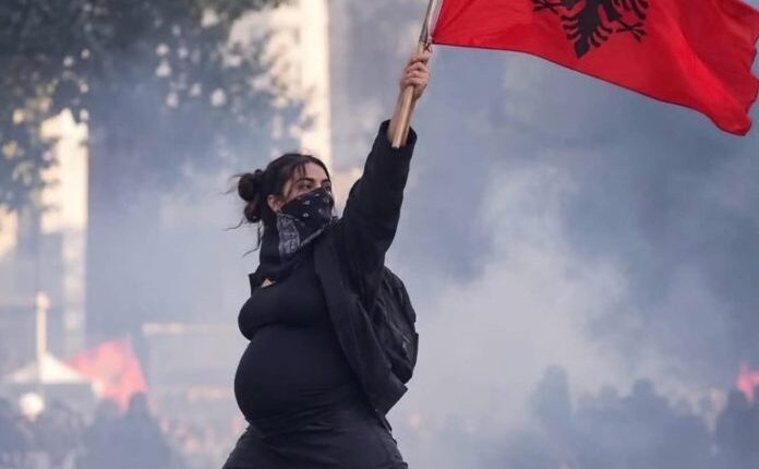Një grua shtatzënë lartëson flamurin kombëtar në mes të tymit dhe protestave në Tiranë